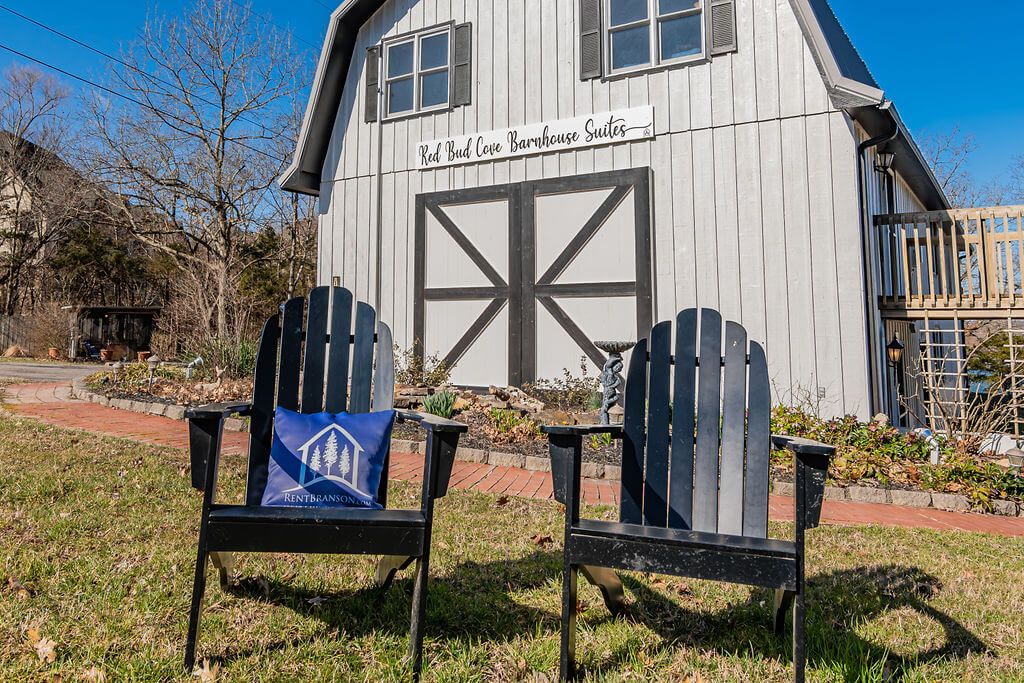 Una casa de campo rústica con una amplia puerta de barnal, dos sillas Adirondack con cojines azules, y un letrero que dice "Red Bud Cove Barnhouse Suites".