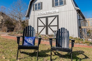 Una casa de campo rústica con una amplia puerta de barnal, dos sillas Adirondack con cojines azules, y un letrero que dice "Red Bud Cove Barnhouse Suites".