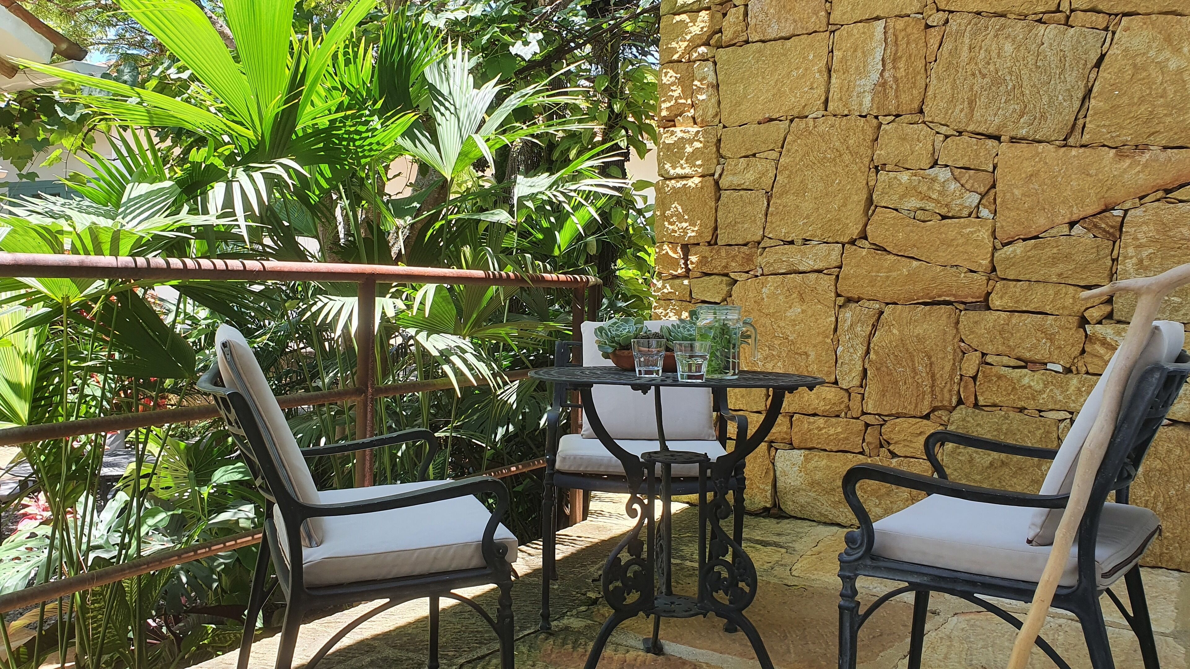 Un patio con un muro de piedra, dos sillas, una mesa con un vaso y abundante follaje.