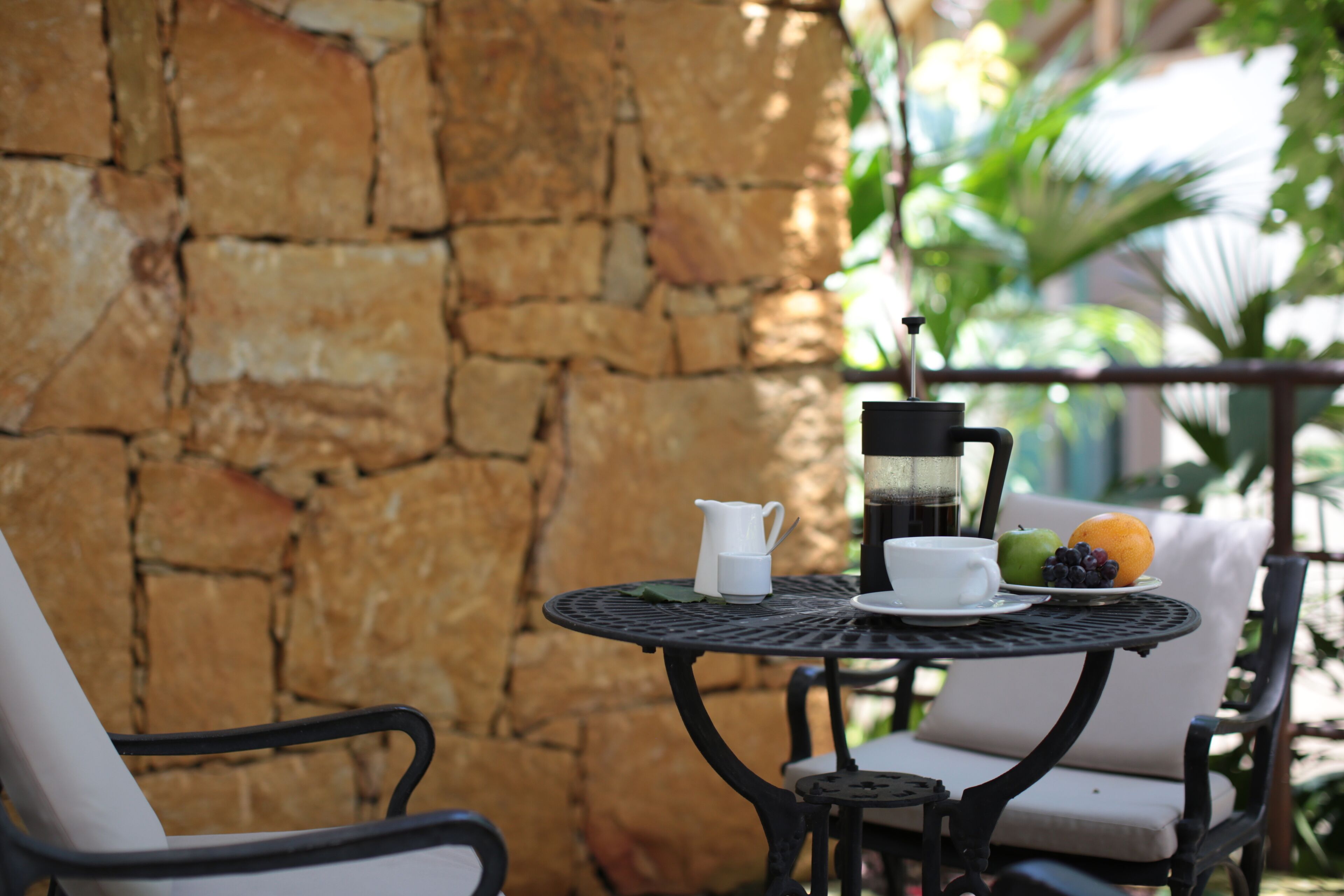A round table with a coffee maker, a white cup, and a plate with fruit. There are two chairs with cushions. The background features a stone wall and greenery.