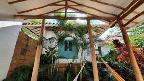 Un espacio al aire libre cubierto con una pérgola de madera, exuberante vegetación tropical y una puerta azul visible entre la estructura.