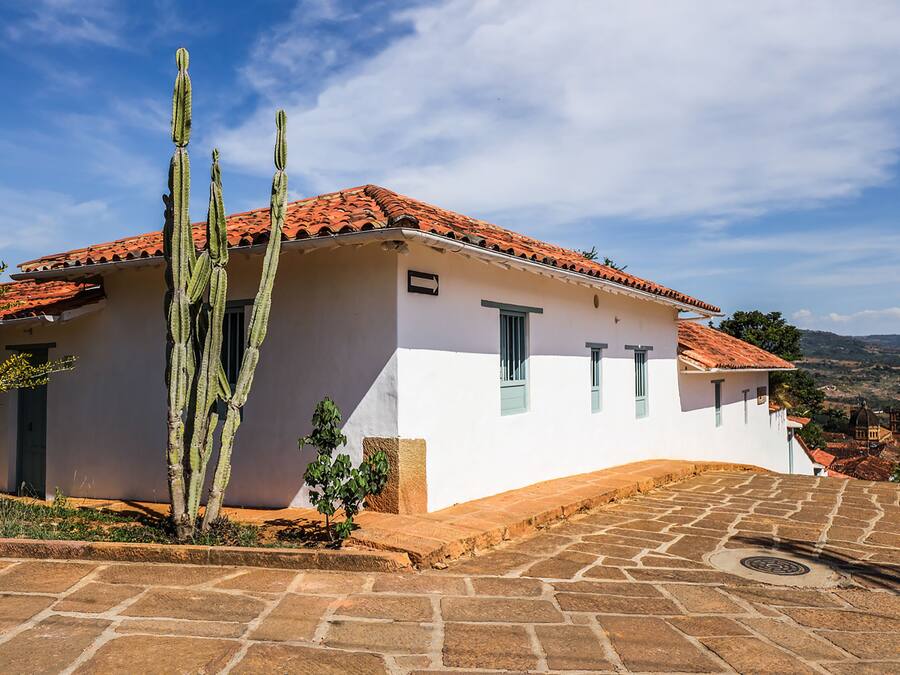 A white building with a red-tiled roof, a cactus in the foreground, and a stone-paved area.