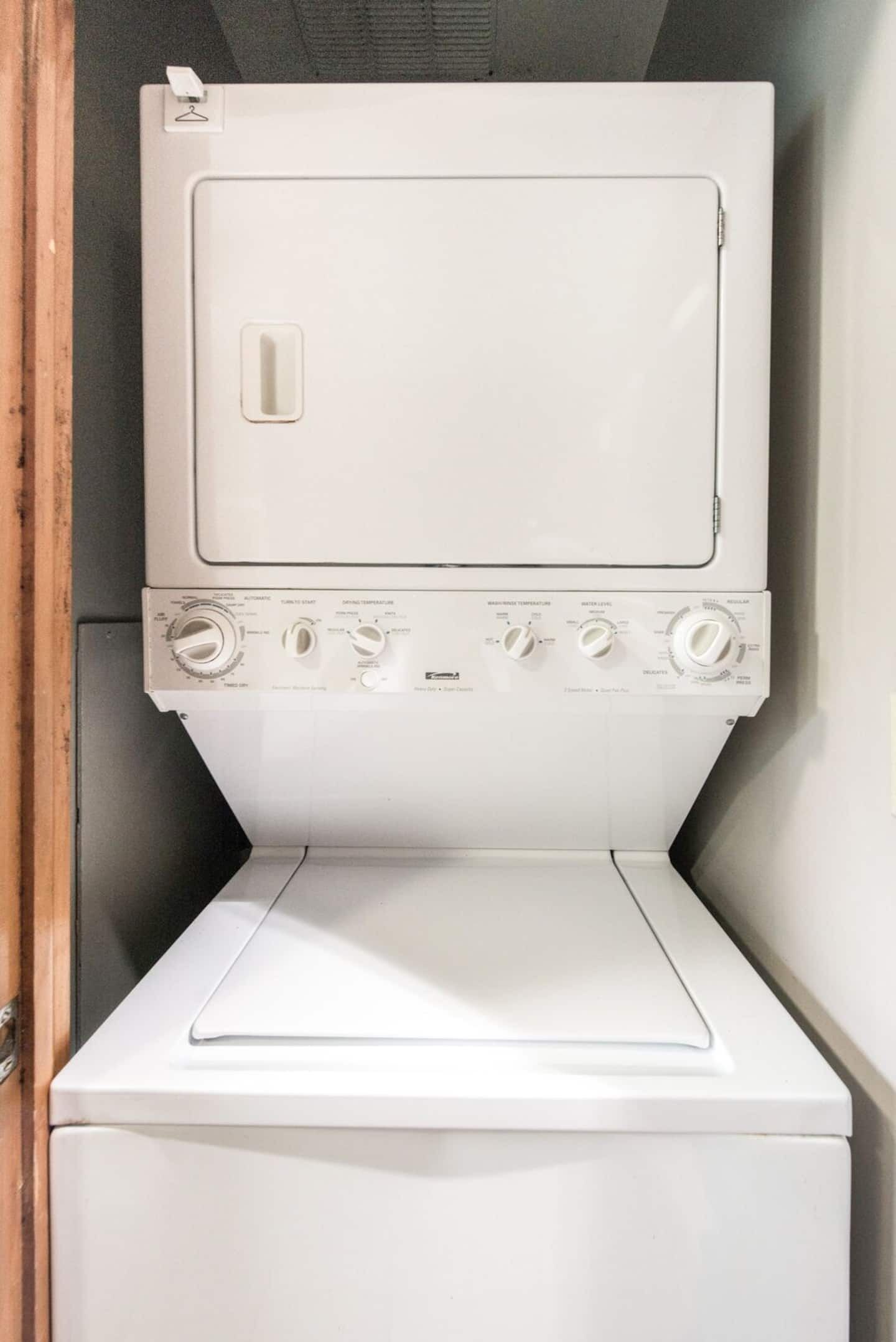 A white washing machine with control knobs and a door, placed in a corner with a wooden door frame.