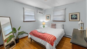 Ein Schlafzimmer mit einem Bett, zwei Fenstern mit Jalousien, einer Deckenleuchte, einem Nachttisch mit Lampe und einem Spiegel.