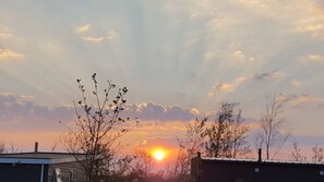 A sunset with the sun partially obscured by clouds, silhouetted trees, and a building with a flat roof.