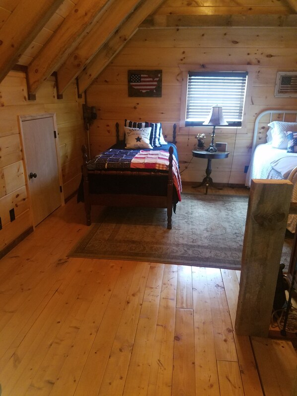 A cozy bedroom with a wooden floor, a bed with a quilt, a nightstand, a lamp, and an American flag decoration.