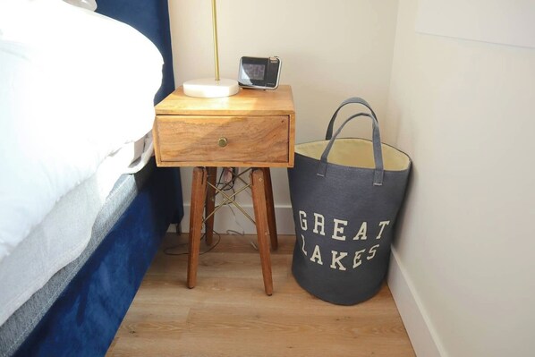 A small wooden bedside table with a digital clock, a grey tote bag with "GREAT LAKES" text, and a bed with blue curtains. - Stunning Downtown Condo: Capri 106 (Traverse City)