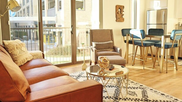 A modern living room with a leather sofa, a round coffee table, and a dining area with blue chairs. - Stunning Downtown Condo: Capri 106 (Traverse City)