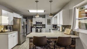 Una cocina moderna con armarios blancos, una isla central con dos taburetes de bar, electrodomésticos de acero inoxidable e iluminación colgante.