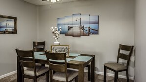 Un comedor con una mesa de madera, cuatro sillas, un ventilador de techo y una obra de arte de varios paneles en la pared.