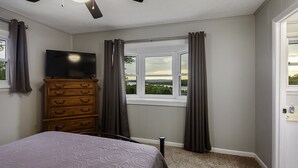 Un dormitorio con una cama, un aparador, un ventilador de techo, una ventana con cortinas y un televisor sobre el aparador.