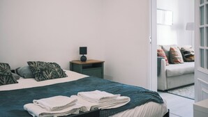 Una cama bien hecha con toallas dobladas, una mesita de noche con lámpara, y vistas a un salón con sofá y cojines.