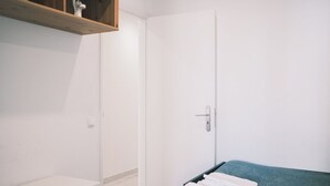 Un dormitorio con un armario blanco, una pequeña estantería con objetos decorativos, una cama con cabecera azul y una mesita de noche blanca.