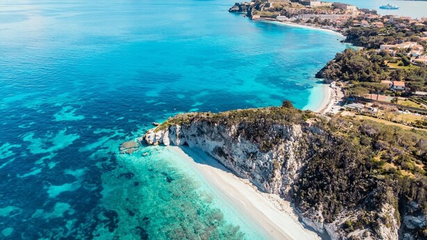An aerial view of a coastal town with clear blue waters, a sandy beach, and a mountainous backdrop.