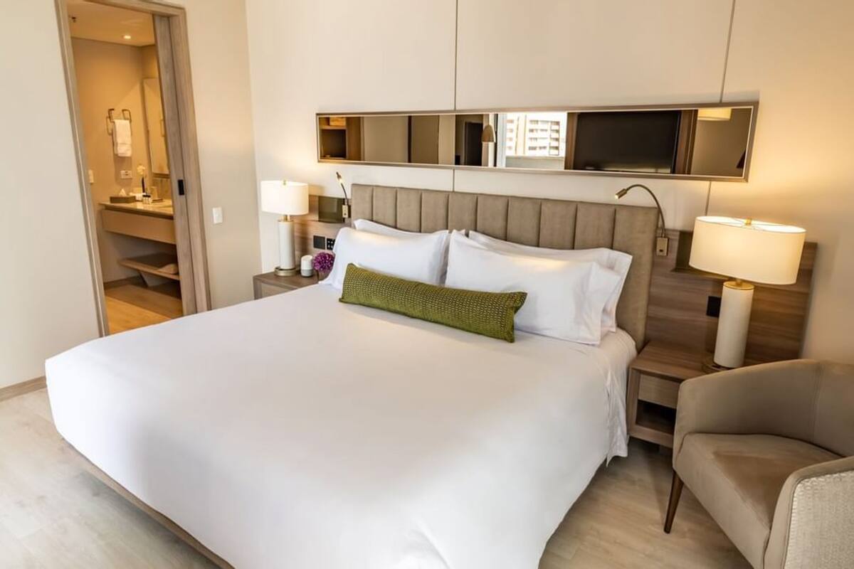 Habitación de hotel con una cama grande, una mesita de noche con lámpara, una silla, un espejo sobre la cama y una puerta que da a un baño.
