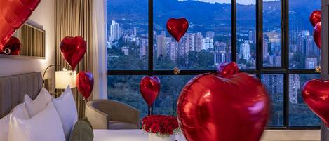 Une chambre d’hôtel avec une grande fenêtre, un lit, une table de chevet ornée d’un vase de roses rouges et des ballons en forme de cœur.