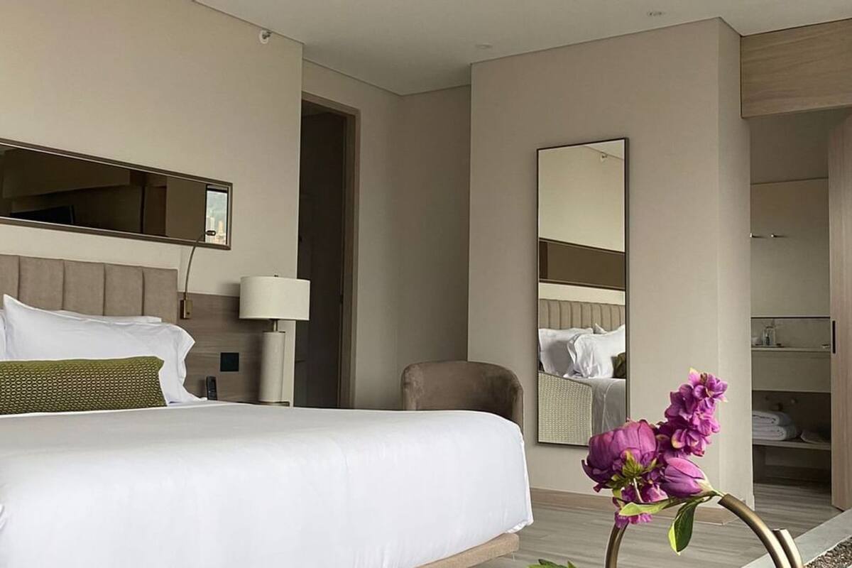Una habitación de hotel con cama, espejo, silla y un jarrón de flores.