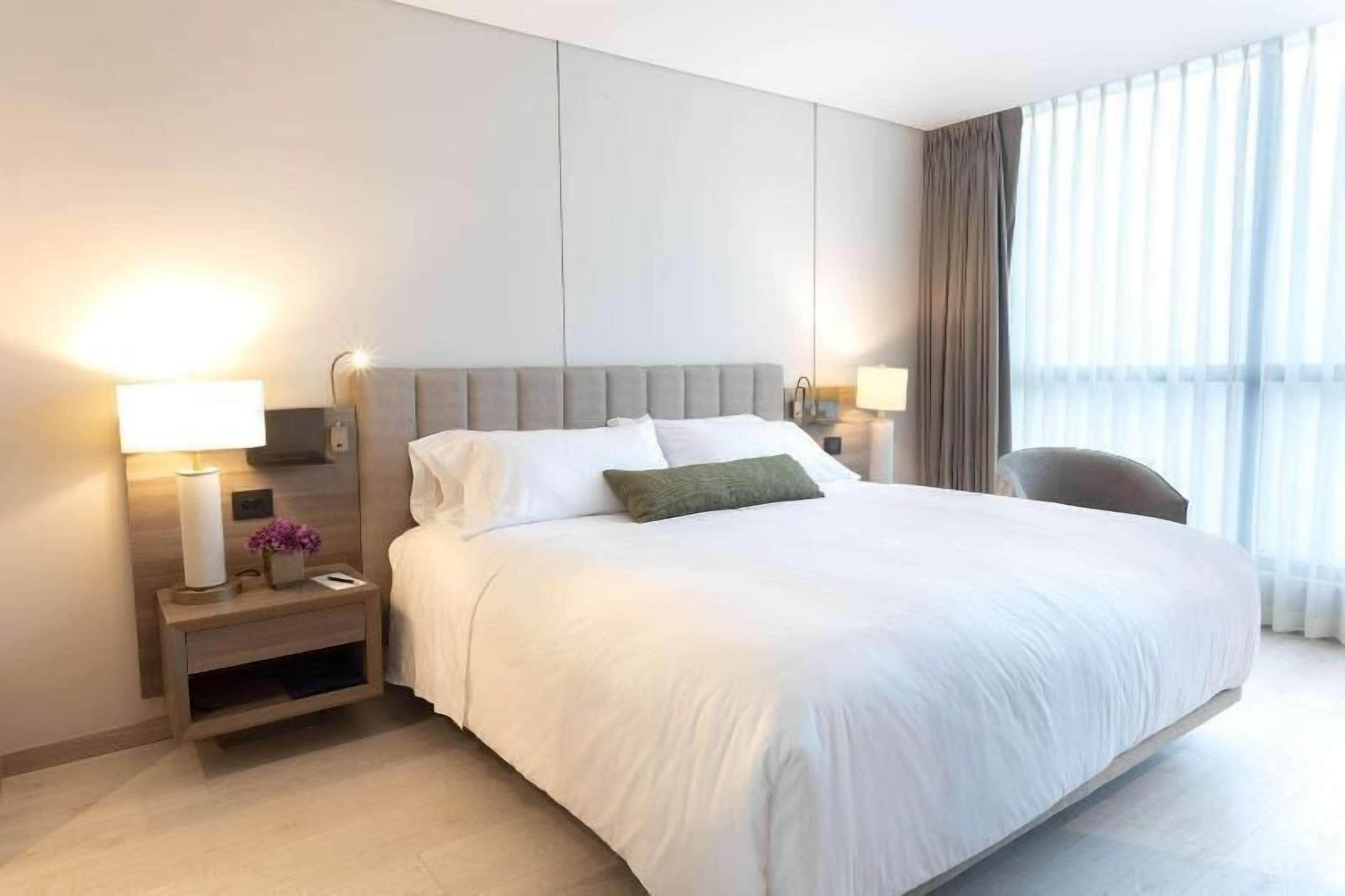 A hotel room with a large bed, two bedside tables with lamps, a chair, and a large window with curtains.