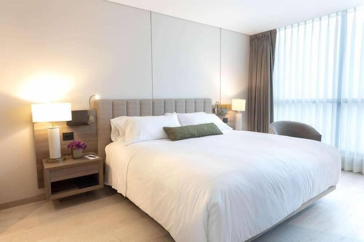 Una habitación de hotel con una cama grande, dos mesitas de noche con lámparas, una silla y un gran ventanal con cortinas.