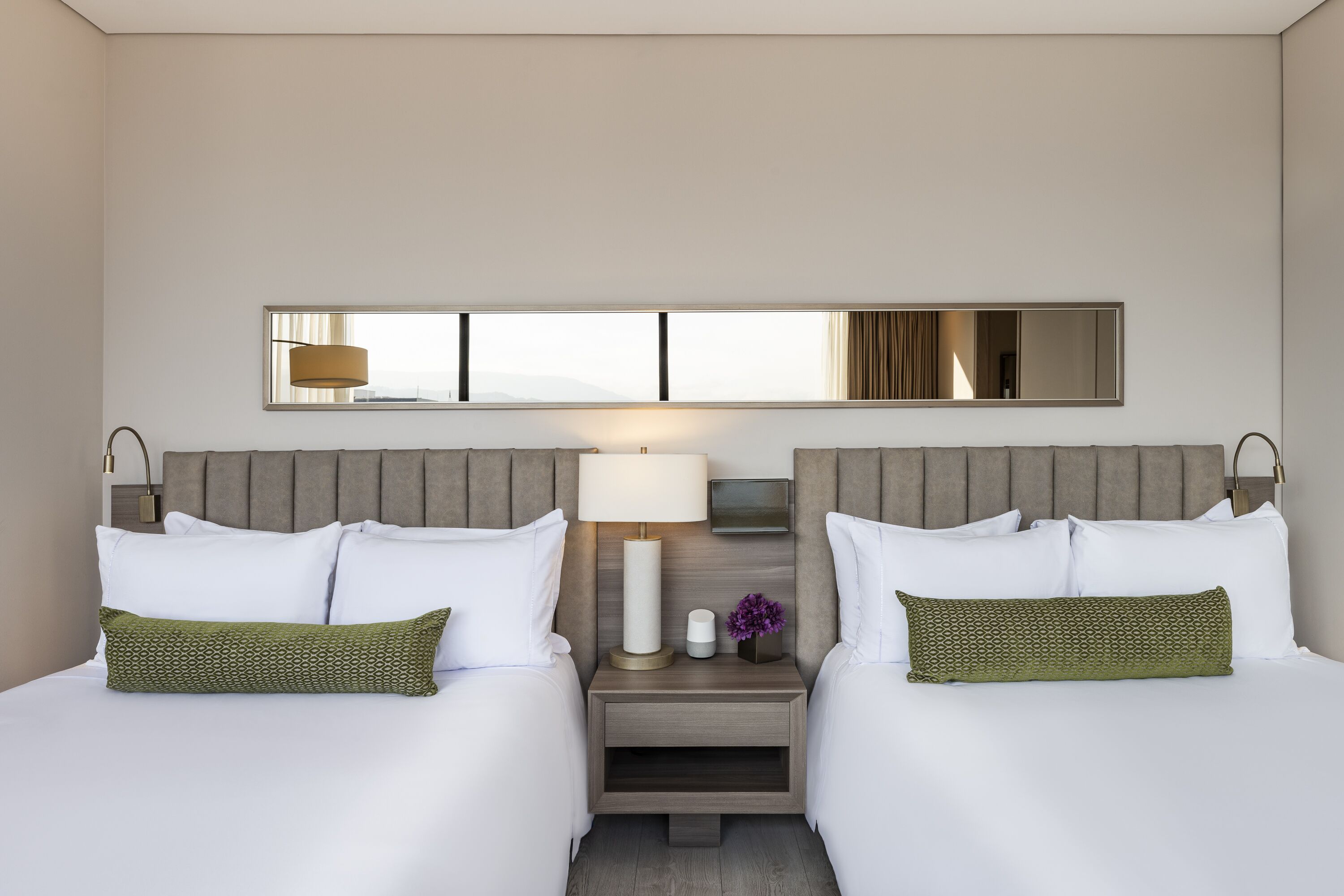 A hotel room with two beds, a nightstand with a lamp, a mirror, and a window with a view.