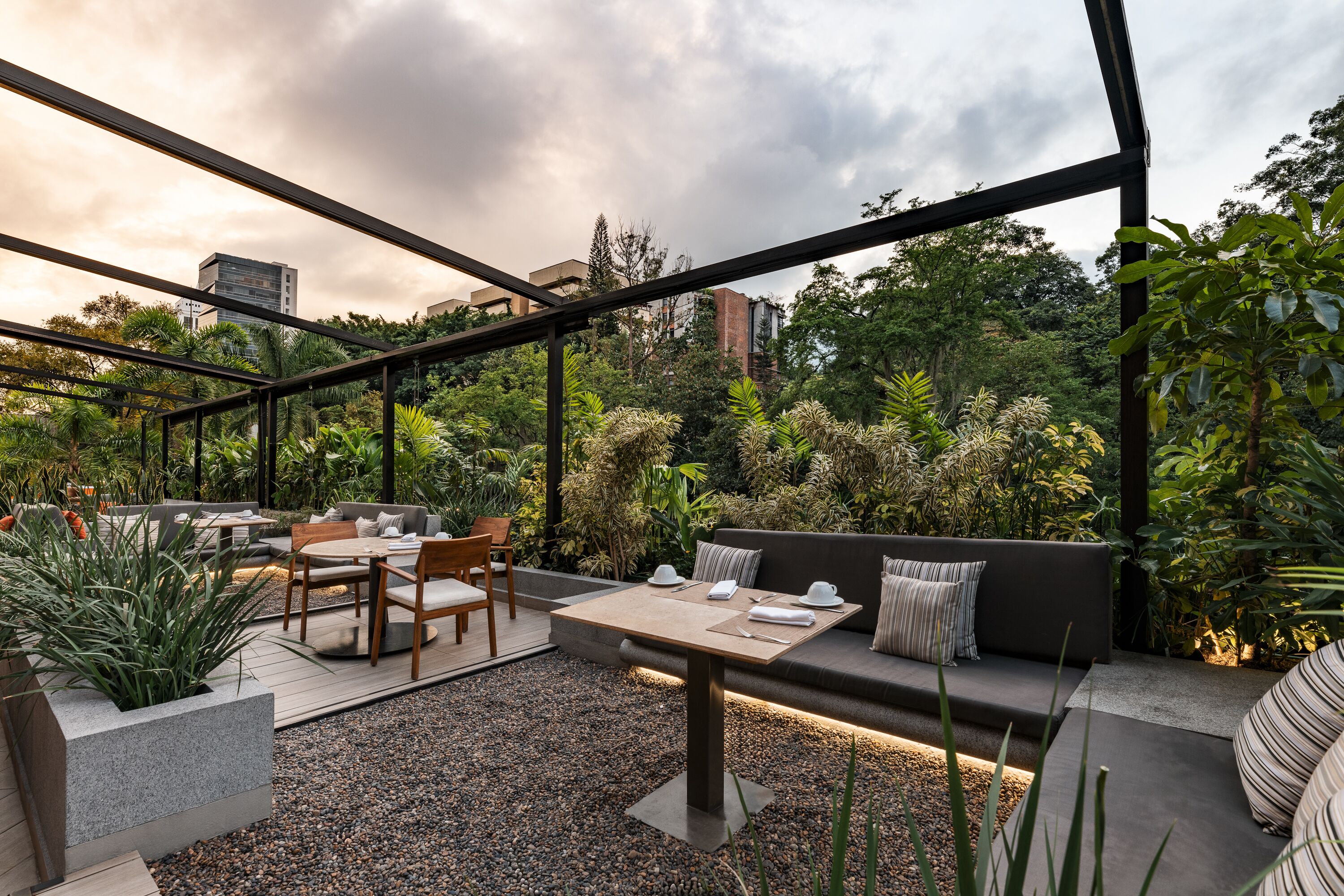 Un patio exterior con una zona de estar moderna, que incluye un sofá y una mesa, rodeado de exuberante follaje y vistas a la ciudad.