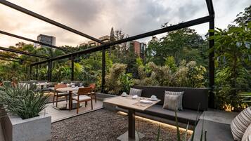 Un patio exterior con una zona de estar moderna, que incluye un sofá y una mesa, rodeado de exuberante follaje y vistas a la ciudad.