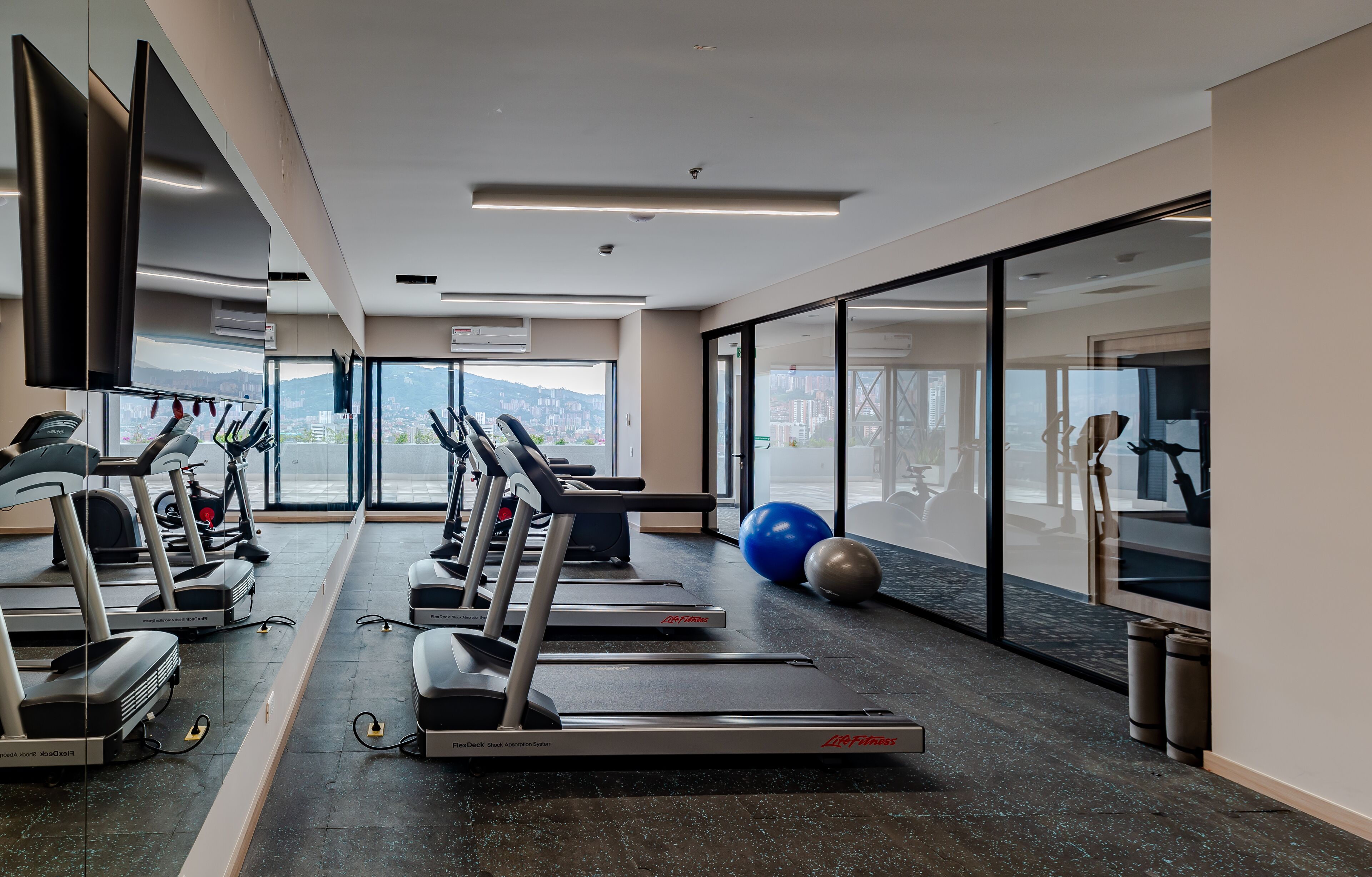 Un gimnasio moderno con cintas de correr, pelotas de ejercicio y un amplio ventanal con vista a la ciudad.