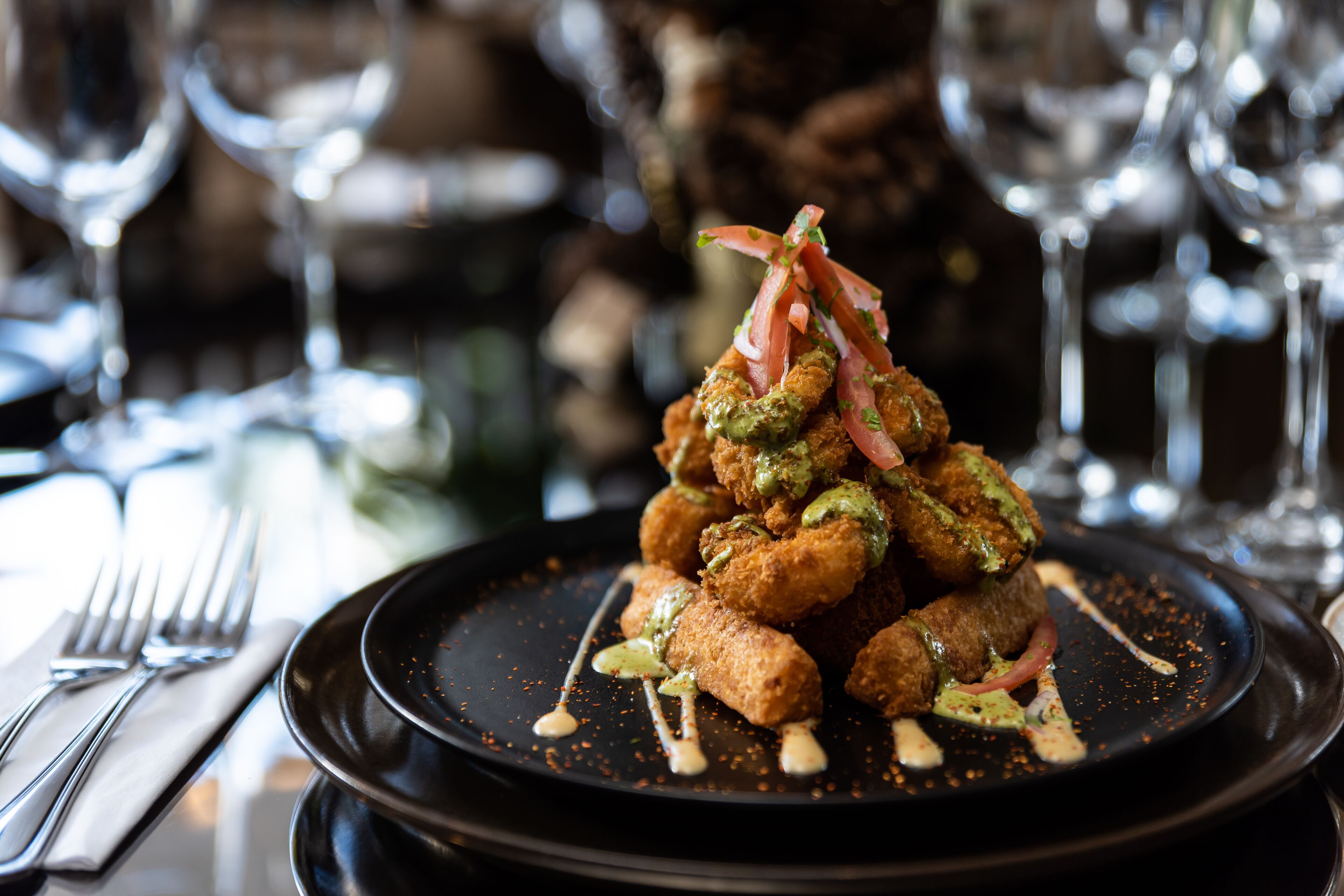 A black plate with a stack of breaded food items, garnished with herbs and sauce, on a table with a napkin and fork.