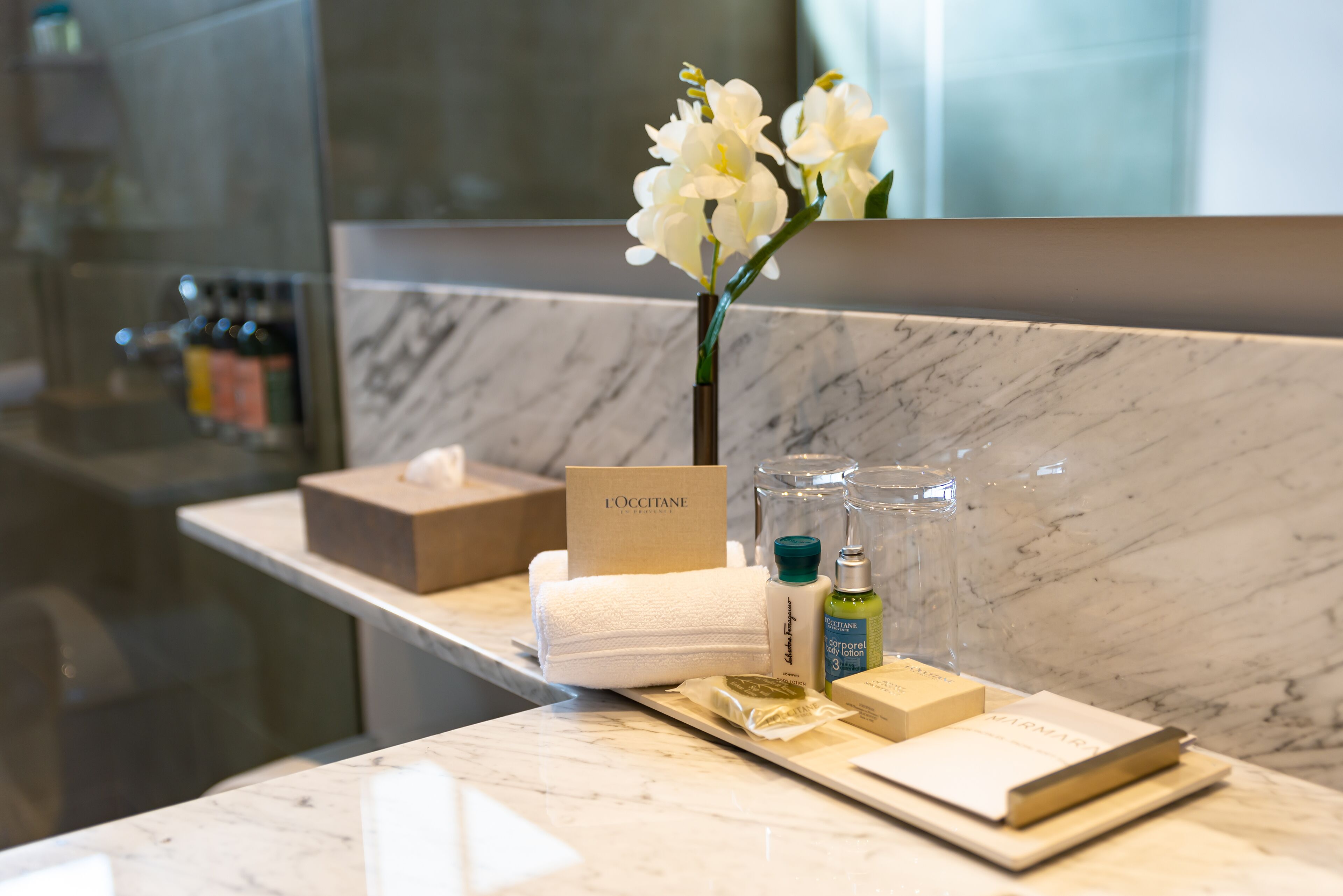 A hotel reception area with a marble counter, a vase of white flowers, toiletries, and a L'OCCITANE box.