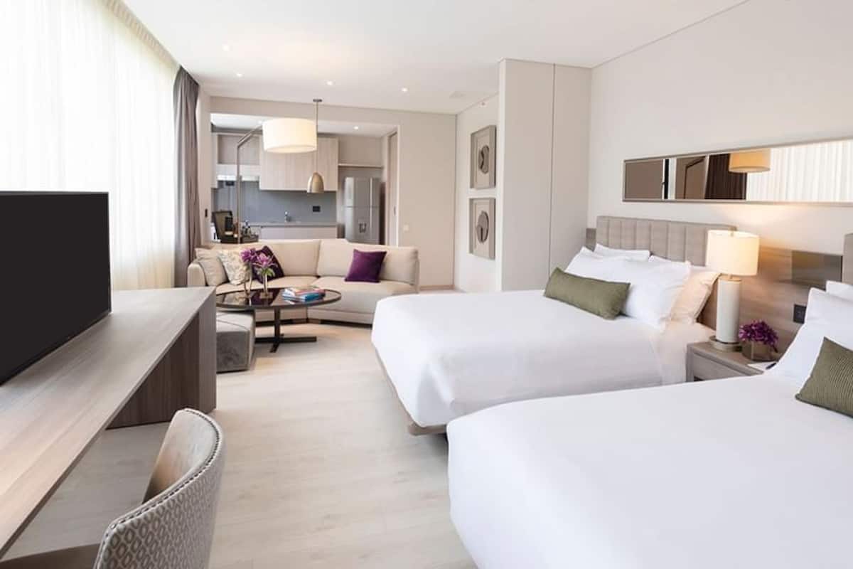 Una habitación de hotel con dos camas, televisión, escritorio, silla, mesa de centro y vista a la cocina.
