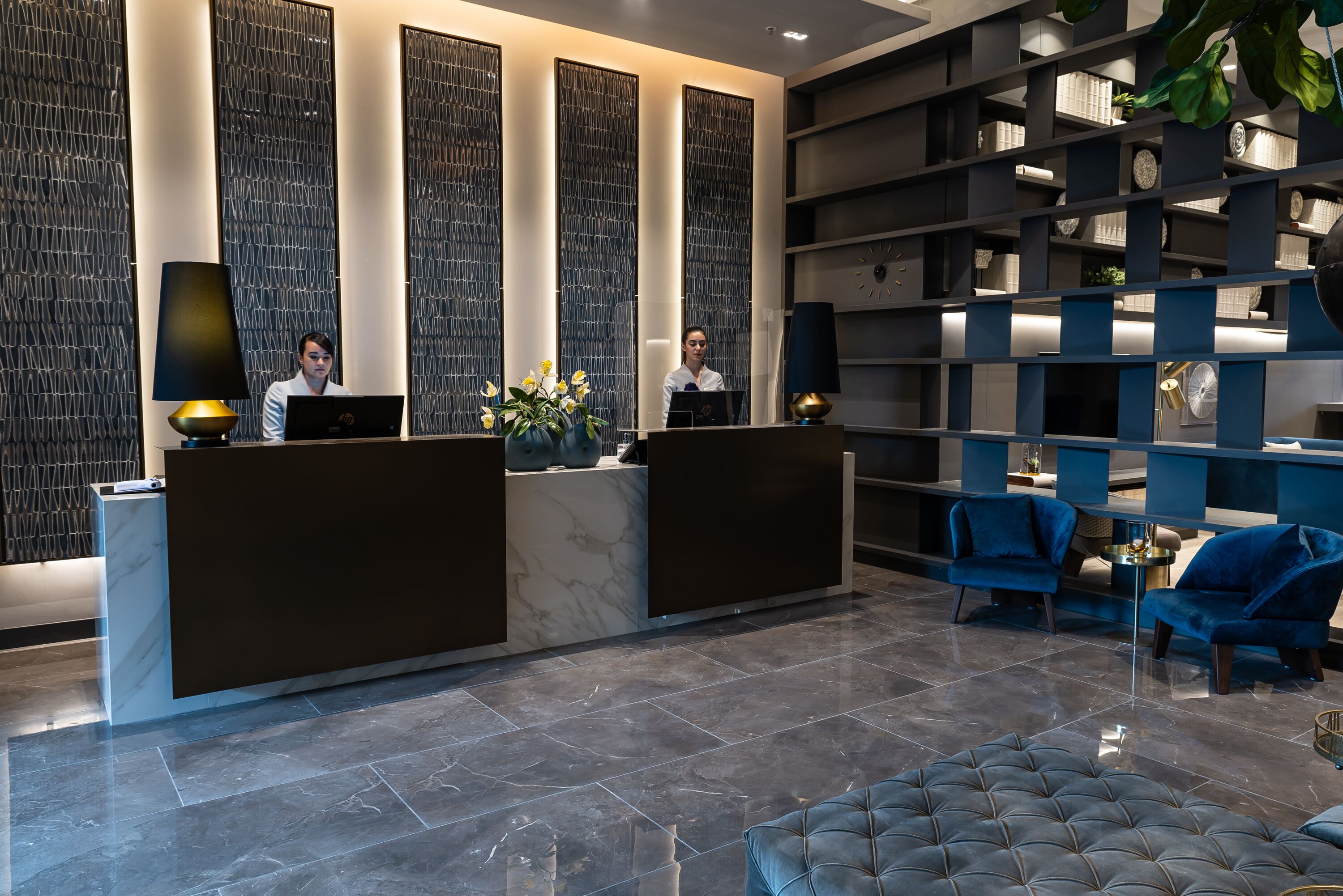 Un'area di accoglienza con un bancone in marmo, due addetti alla reception, un divano blu e una libreria con oggetti decorativi.