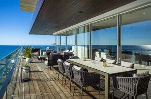 Un área de comedor exterior moderna con vista al océano, que incluye una mesa larga con sillas y plantas en macetas.