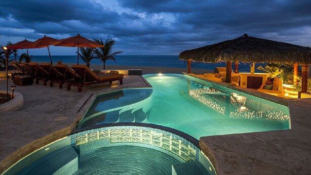 A pool area with lounge chairs and umbrellas, a thatched-roof structure, and a view of the ocean.