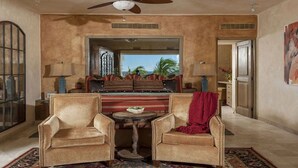 A spacious living room with a high ceiling, a ceiling fan, and a view of the outdoors through a large window. There are two armchairs, a small round table, and a sofa with a red throw blanket.