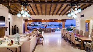 A spacious living area with a large sofa, dining table, and chairs, a TV, and a view of a pool and ocean at dusk. - Beautiful Holiday Villa in a Prime Location in Cabo San Lucas 1007 (Cabo San Lucas)