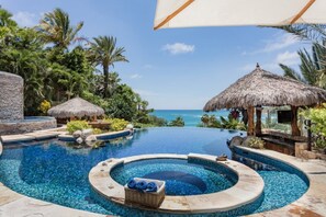 A pool area with a view of the ocean, surrounded by tropical vegetation and thatched-roof structures.