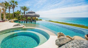 Une piscine à débordement offrant une vue imprenable sur l’océan, entourée de rochers et de palmiers.