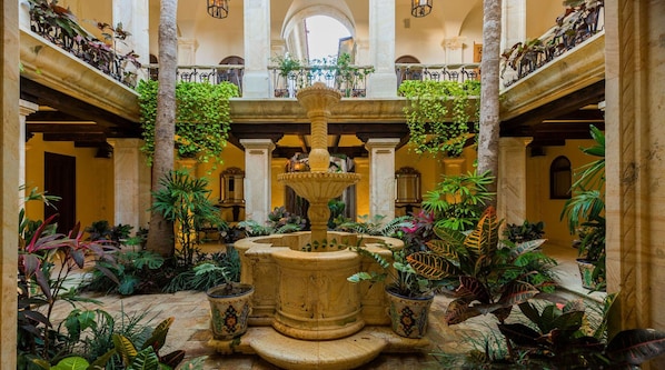 Une cour intérieure avec une fontaine centrale, entourée d’une végétation tropicale luxuriante et de colonnes.