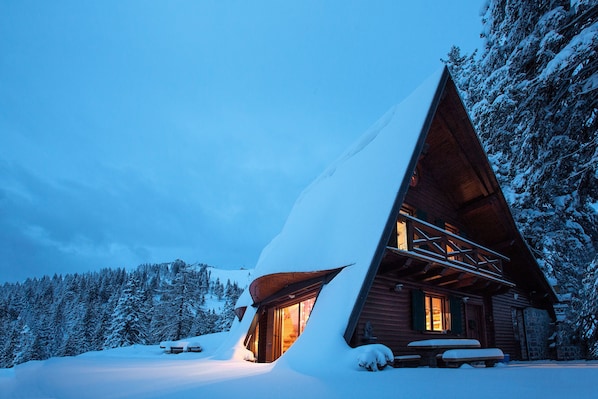 Una cabaña nevada con techo inclinado, fachada de madera y un interior iluminado.