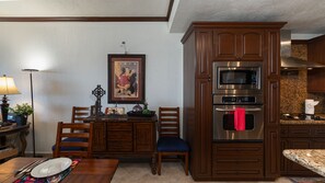 Een keuken met houten kastjes, een RVS magnetron en een oven. Er is een eettafel met stoelen en een lijst met een foto aan de muur.