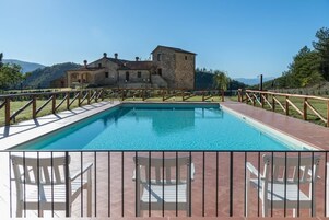 A swimming pool with a wooden deck and railings, surrounded by a stone building with a view of hills.