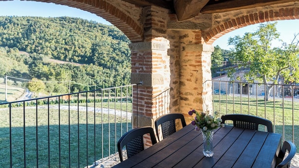 Een overdekt terras met een tafel en stoelen, een bakstenen muur en uitzicht op een glooiend landschap.