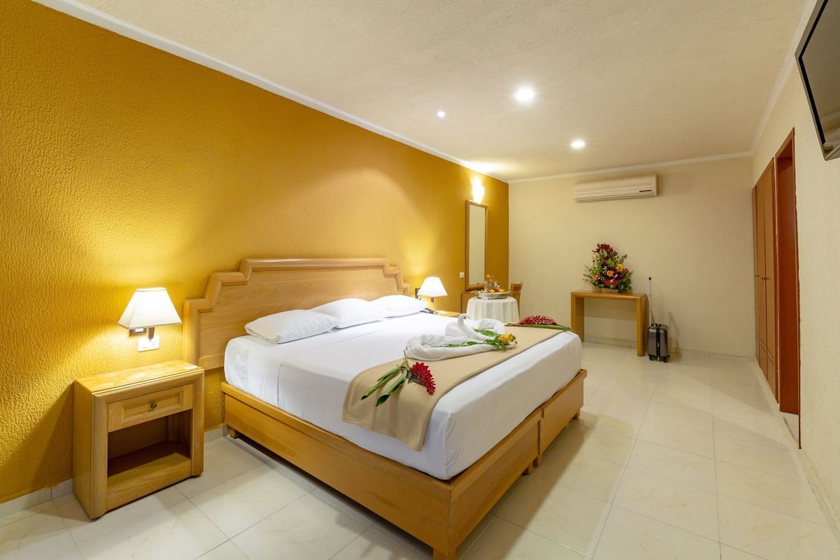 A hotel room with a wooden bed, bedside lamps, a small table with a vase of flowers, an air conditioning unit, and a wardrobe.