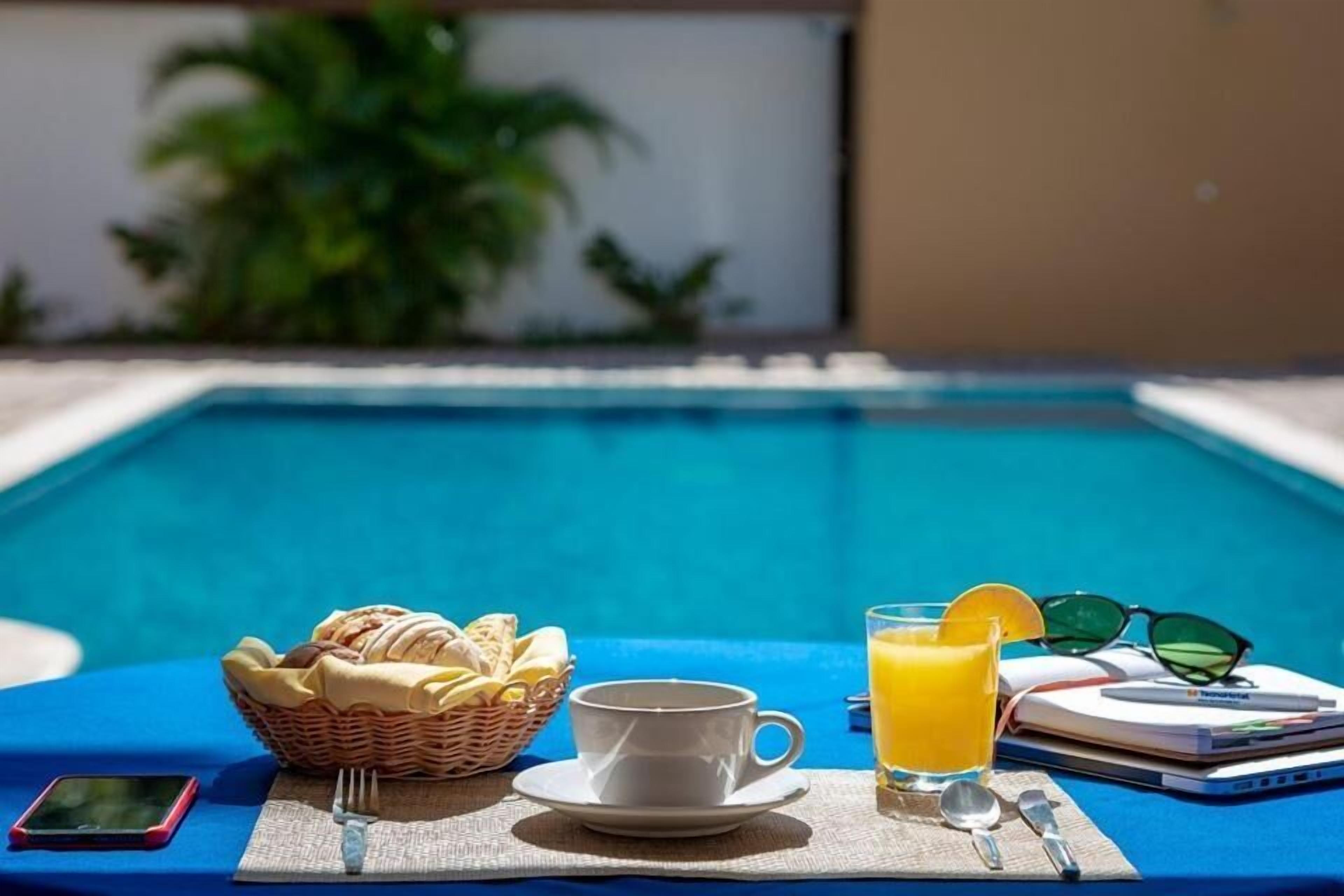 Desayuno junto a la piscina con una cesta de pasteles, una taza de café, un vaso de zumo de naranja y un teléfono inteligente sobre un mantel azul.