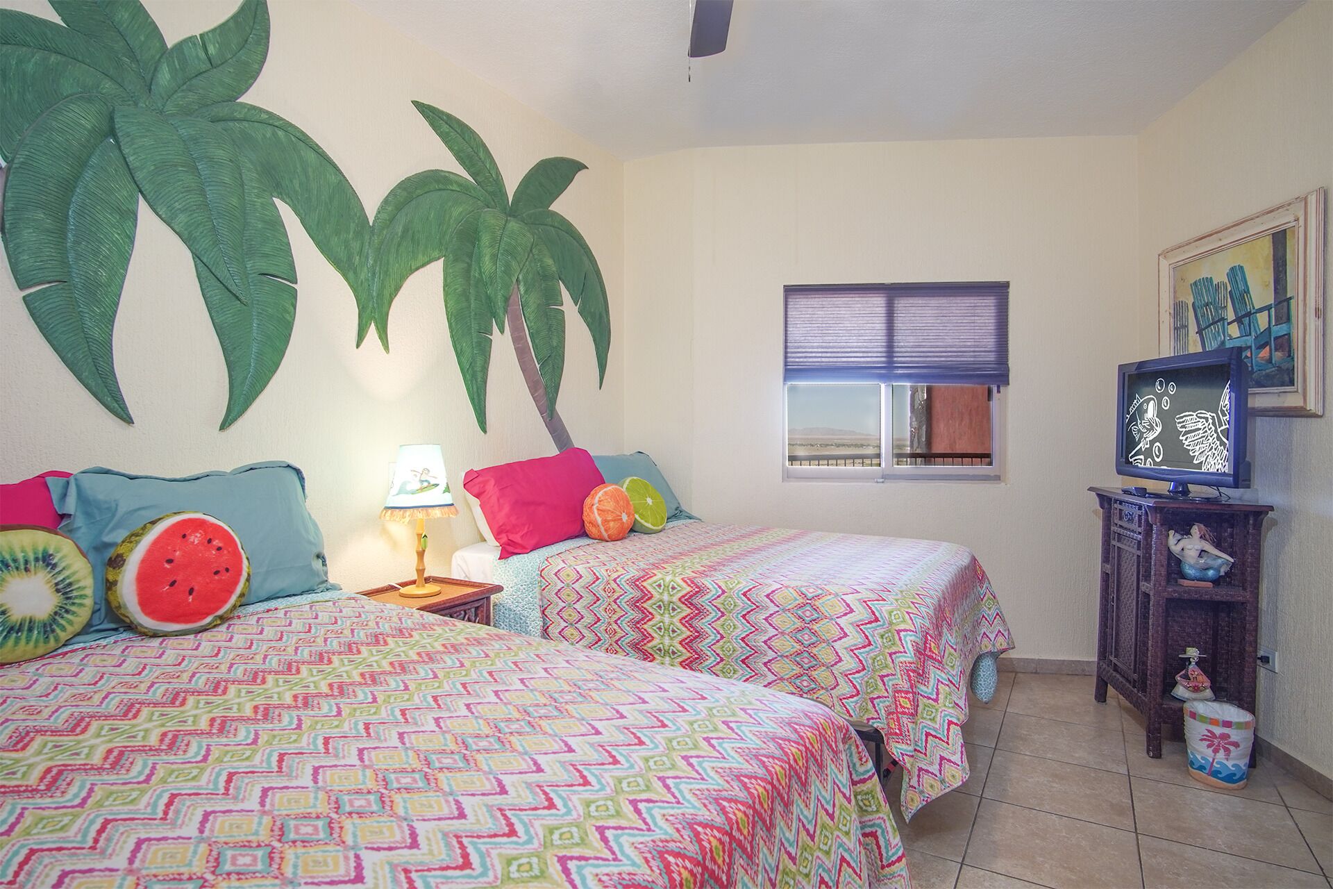 Un dormitorio con dos camas, un televisor, una ventana con persianas y un mural de palmeras.