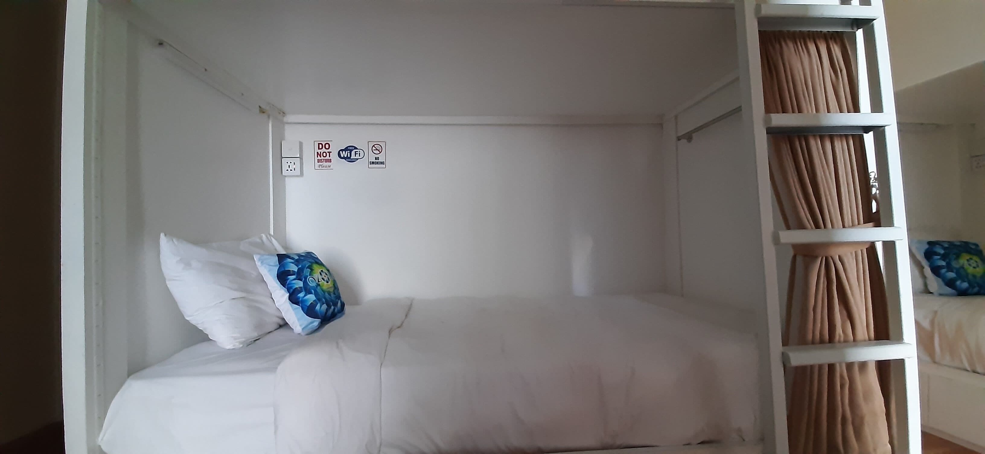 Una habitación pequeña con cama individual, ropa de cama blanca, almohada azul y blanca y cabecera blanca. Hay tres letreros en la pared: “NO USAR”, “NO WIFI” y “NO ENTRAR”.