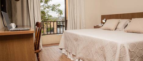 A hotel room with a bed, a desk with a laptop, a chair, a nightstand with a lamp, and a view of the outside.