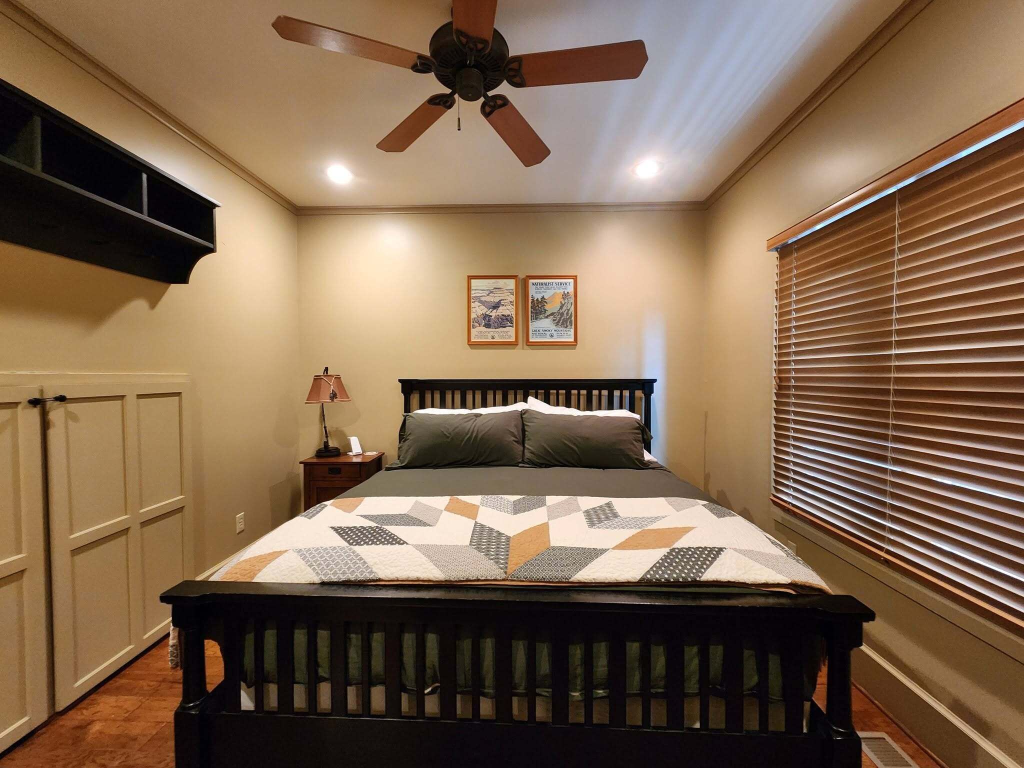 Un dormitorio con cama, ventilador de techo, mesita de noche, lámpara y dos cuadros en la pared.