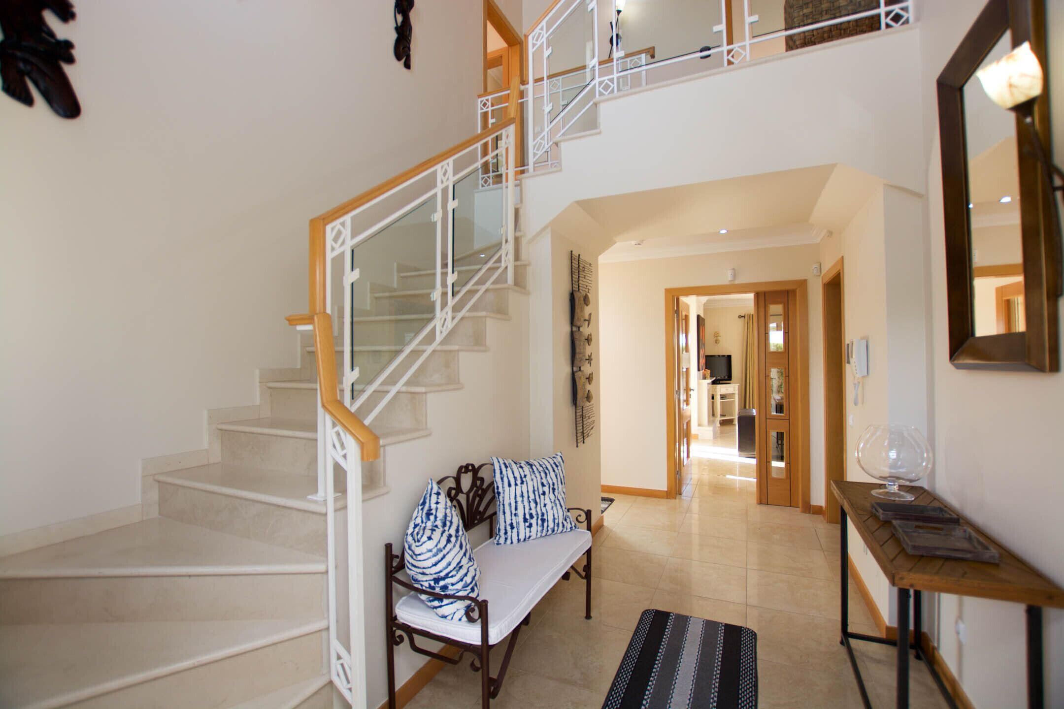 Un vestíbulo amplio con una escalera que conduce a un nivel superior, una zona de estar con un banco y cojines, y una mesa con un jarrón de cristal.