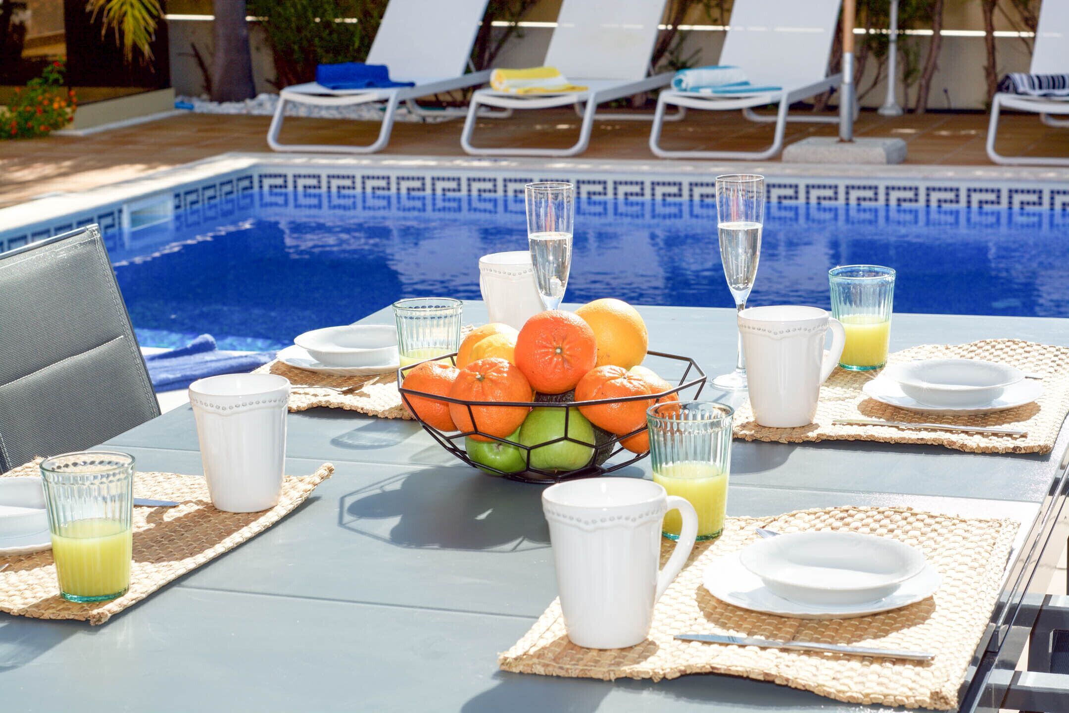 A poolside dining setup with a table set for two, featuring a fruit bowl, glasses, and plates.